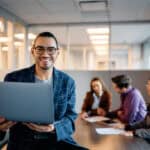 Happy Hispanic businessman using laptop during meeting with coworkers in office.