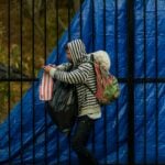A man wearing a hoodie walks with bags by a blue tarp-covered fence on a rainy day