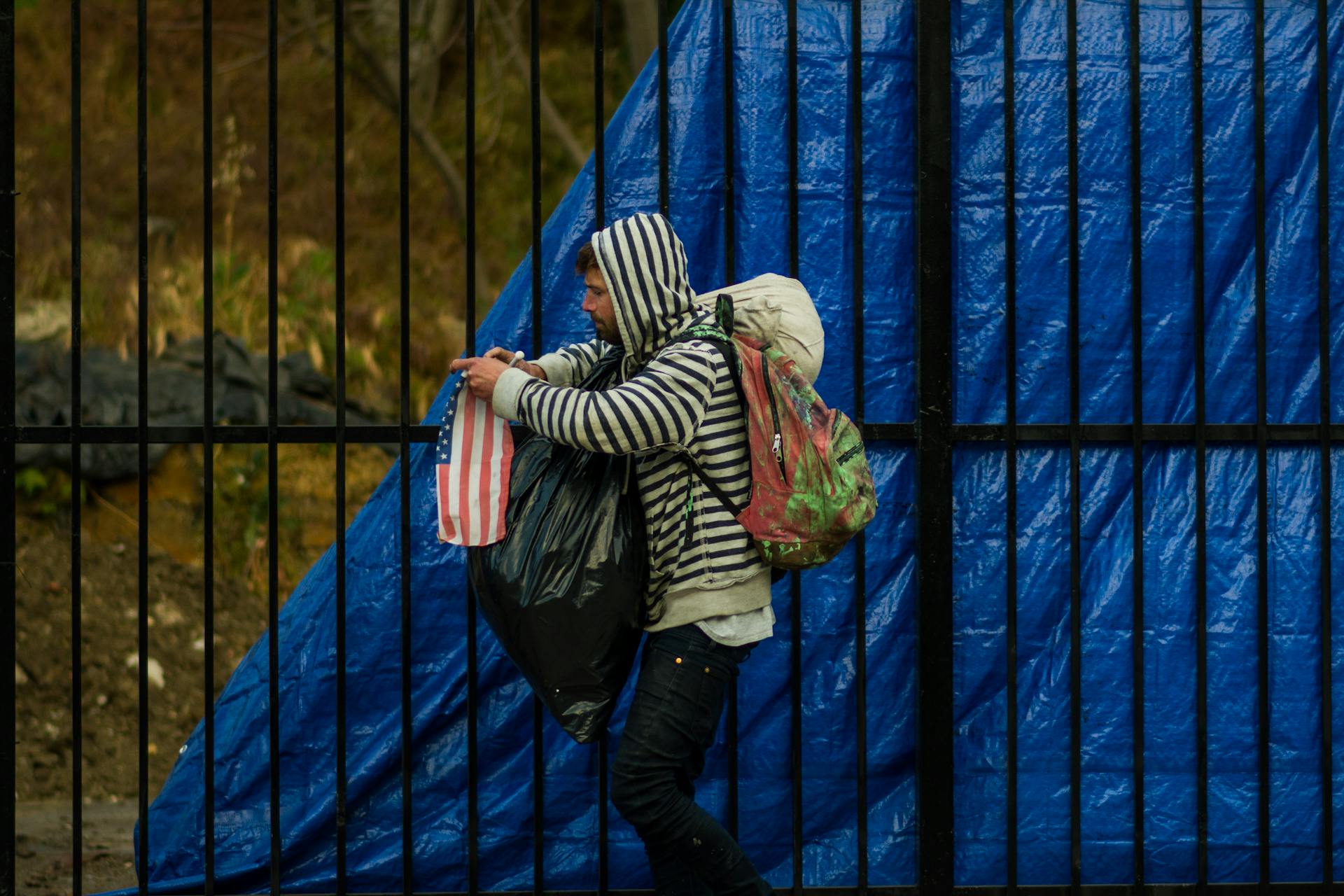 A man wearing a hoodie walks with bags by a blue tarp-covered fence on a rainy day