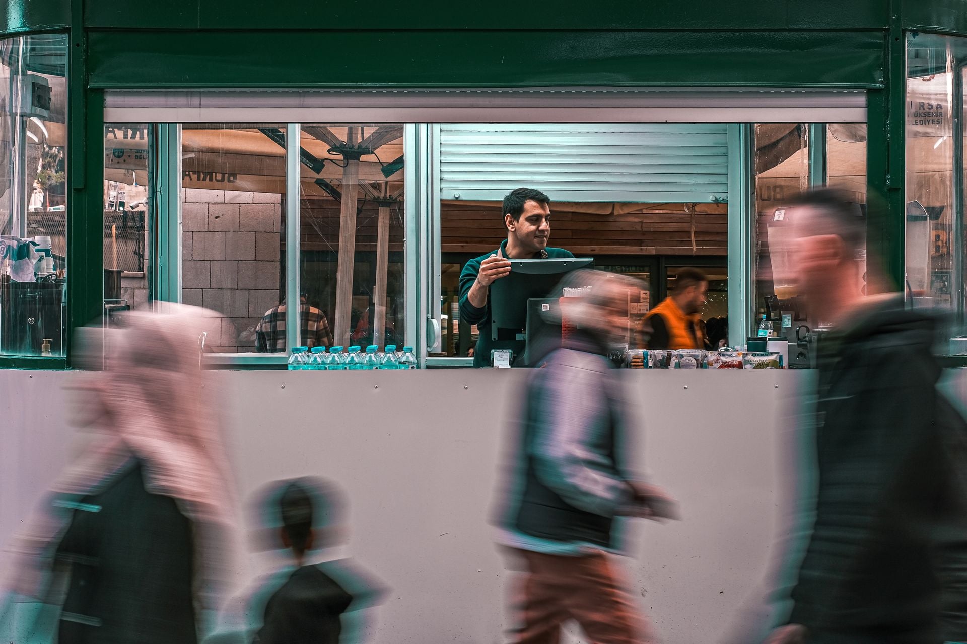 A street vendor serving customers through a kiosk window while people walk by in a busy market setting