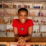 A cheerful woman stands behind a wooden counter, smiling in front of stocked shelves in a store