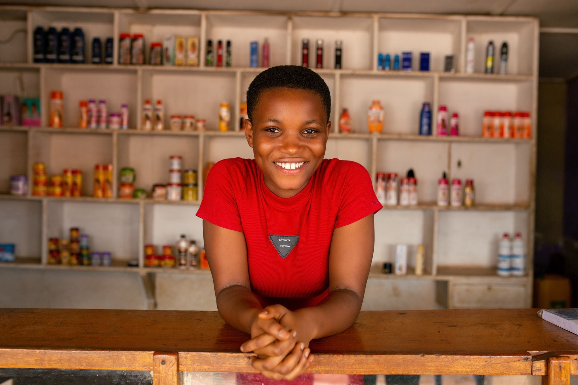A cheerful woman stands behind a wooden counter, smiling in front of stocked shelves in a store