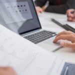 How AI Is Helping Businesses Improve Workplace Training and Documentation 8 Close-up of hands working on documents and a laptop in an office setting, illustrating teamwork and productivity