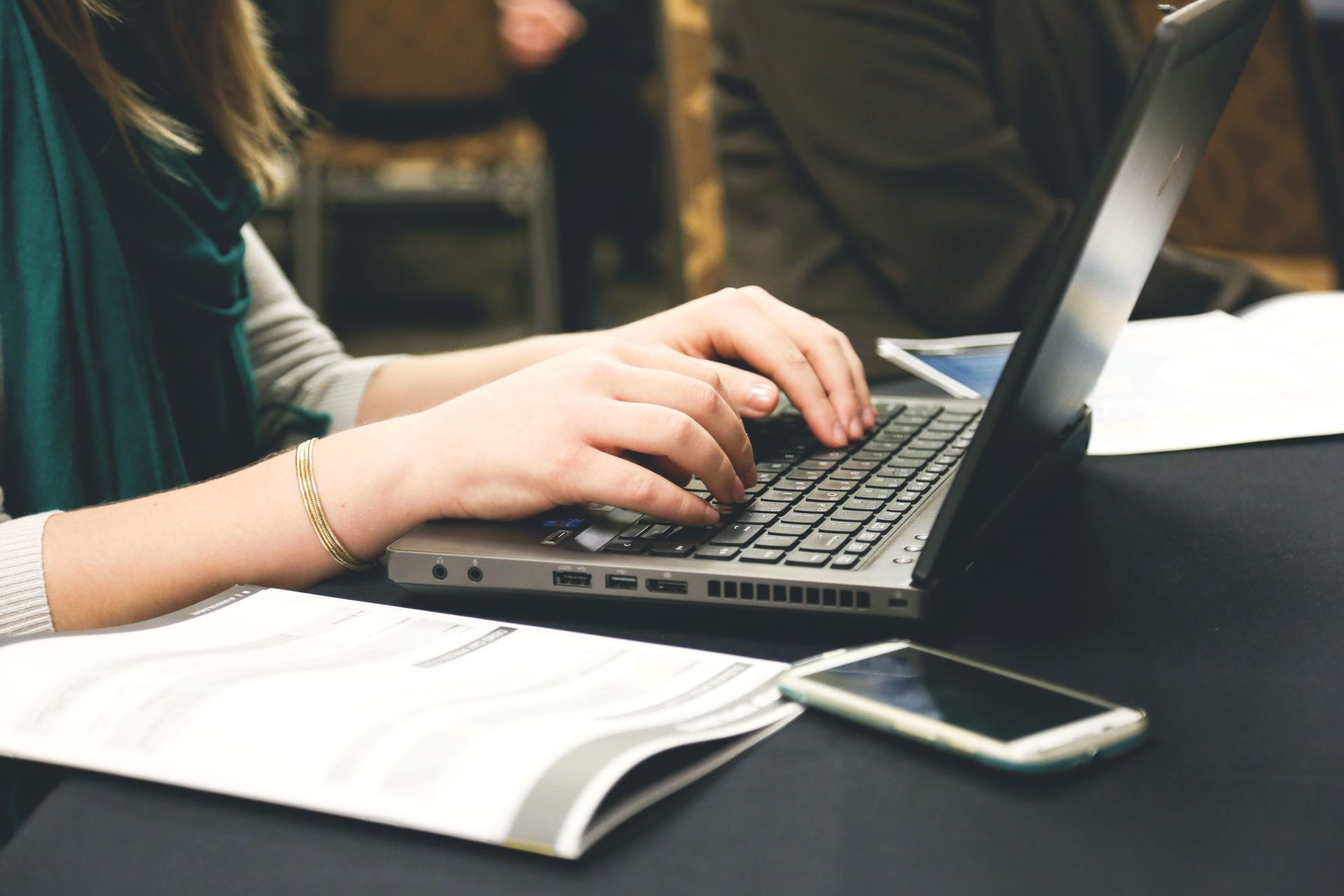 Adult woman typing on a laptop at a desk with a smartphone and documents