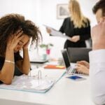 African American woman stressed at office desk with colleagues nearby, conveying workplace pressure