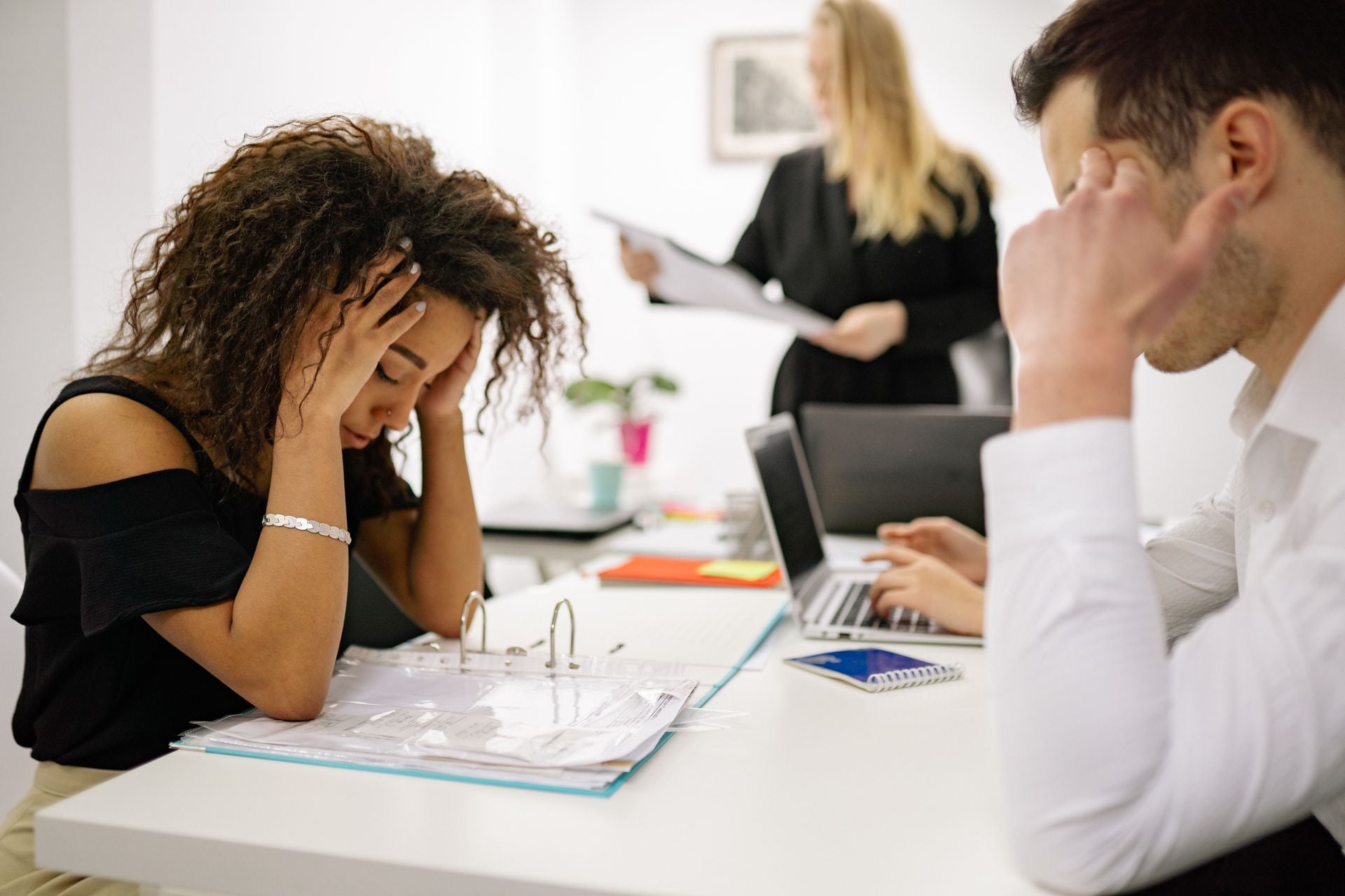 African American woman stressed at office desk with colleagues nearby, conveying workplace pressure